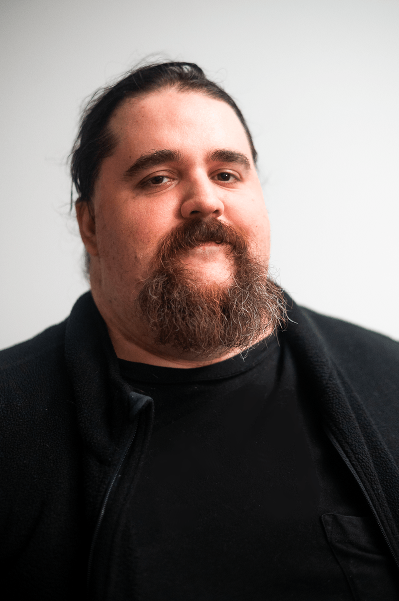 A broad shouldered man with a beard with copper and streaks of white in a shirt and vest